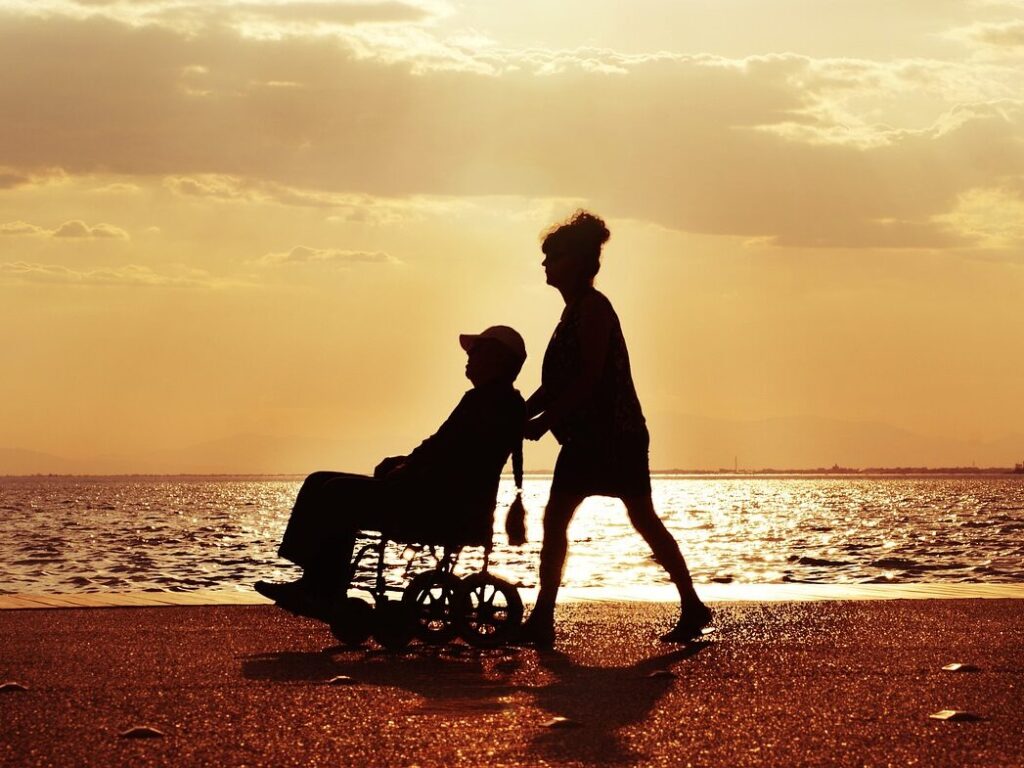 Senior woman pushing her husband in a wheelchair along the beach at sunset, symbolizing compassionate home care services in San Francisco